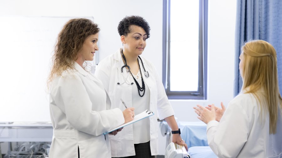 Three female Doctor of Nursing Practice (DNP) program members are discussing a patient's plans. They are all wearing white coats and are in a clinic.